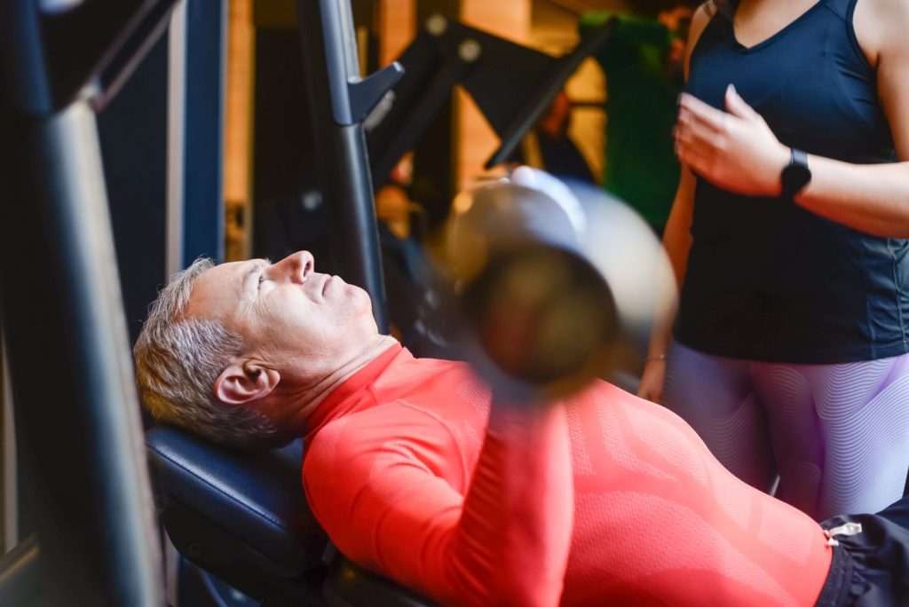 Older adult lifting a barbell under close supervision, progressing from rehabilitation into strength training